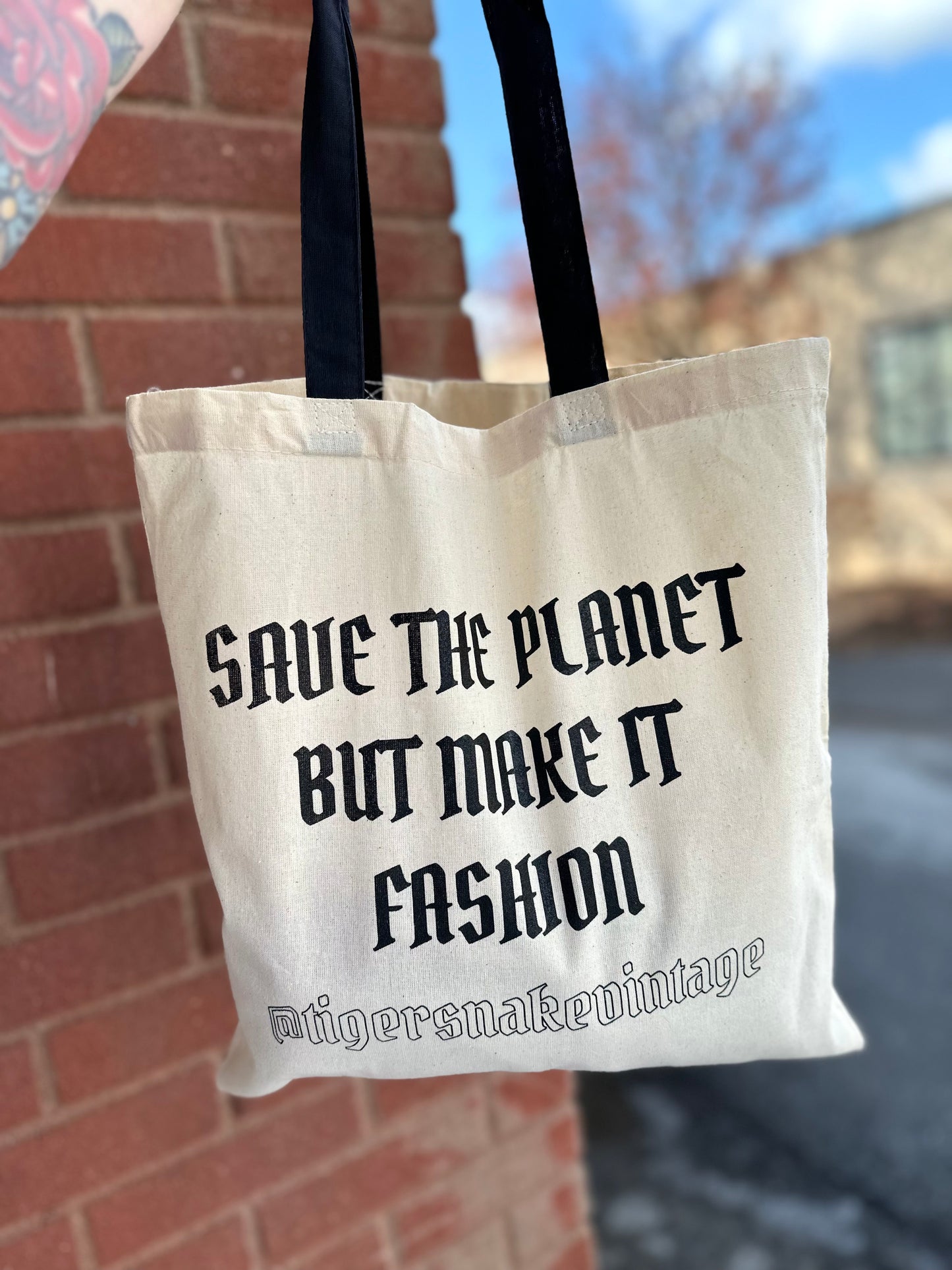 A natural colored canvas tote with black straps printed with "save the planet but make it fashion" along with the instagram handle for Tiger Snake Vintage