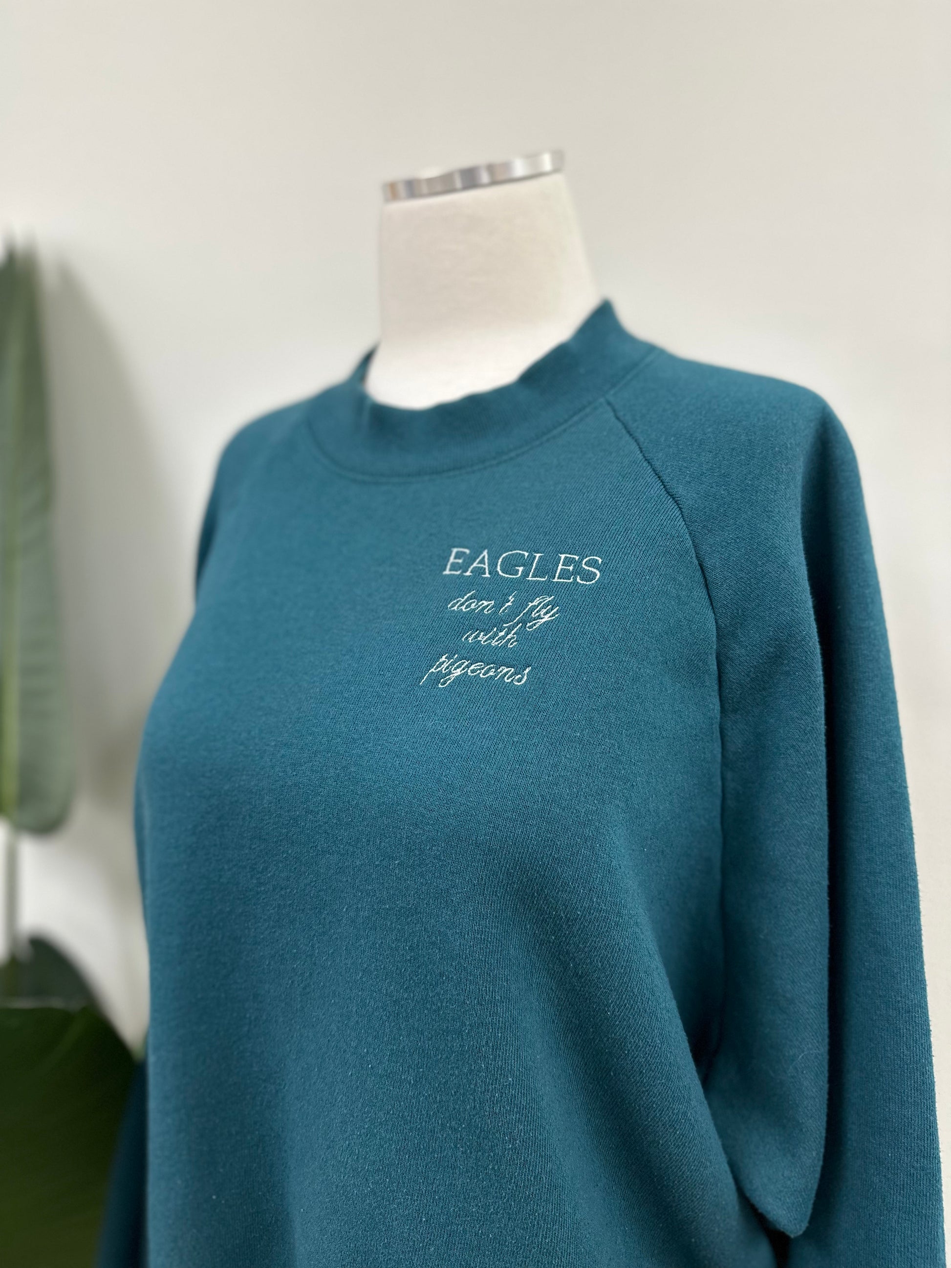 Close up view of a dark teal vintage crewneck with a white embroidery that says "Eagles Don't Fly With Pigeons", a quote said by Real Housewives of New York's Dorinda Medley